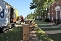 A person pushes a dolly up a ramp into the back of a moving truck with moving boxes lined up along a sidewalk