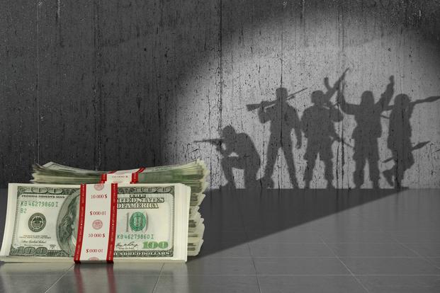 Stacks of 100 dollar bills in the foreground with shadows of troops cast on a wall